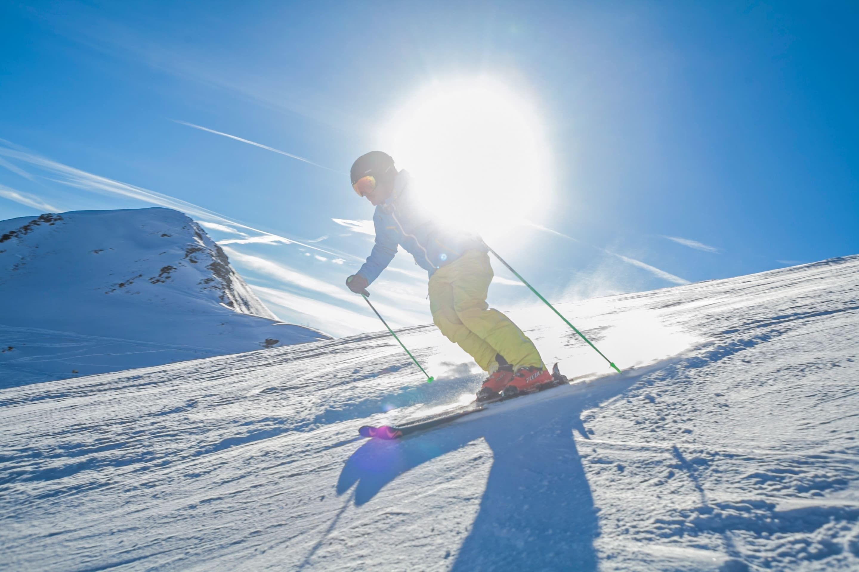 Une personne fait du ski sur une pente enneigée, avec un ciel dégagé et le soleil en arrière-plan.