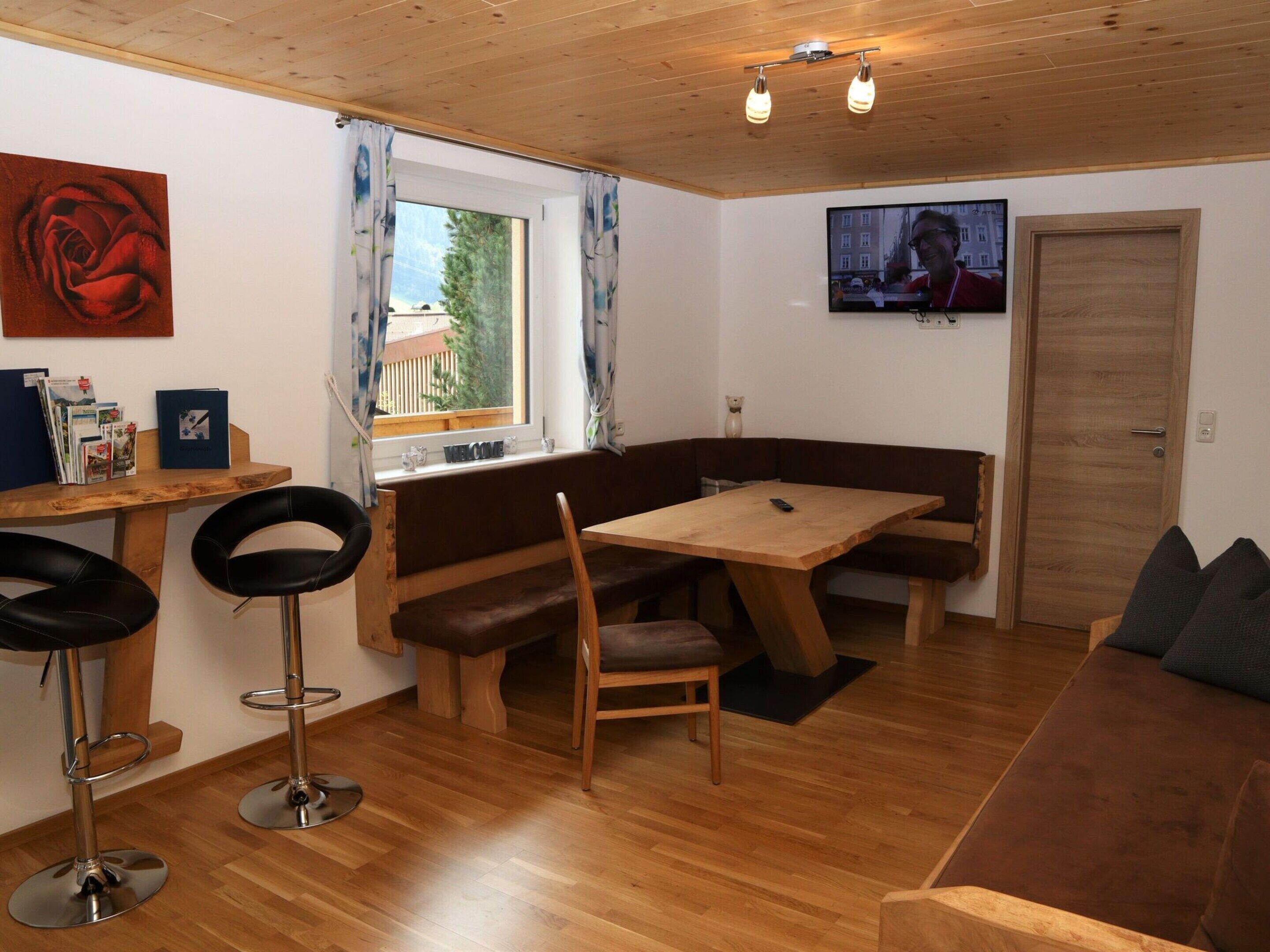 Une pièce chaleureuse avec une table en bois, des bancs et un canapé. Il y a un téléviseur accroché au mur et une fenêtre avec des rideaux.