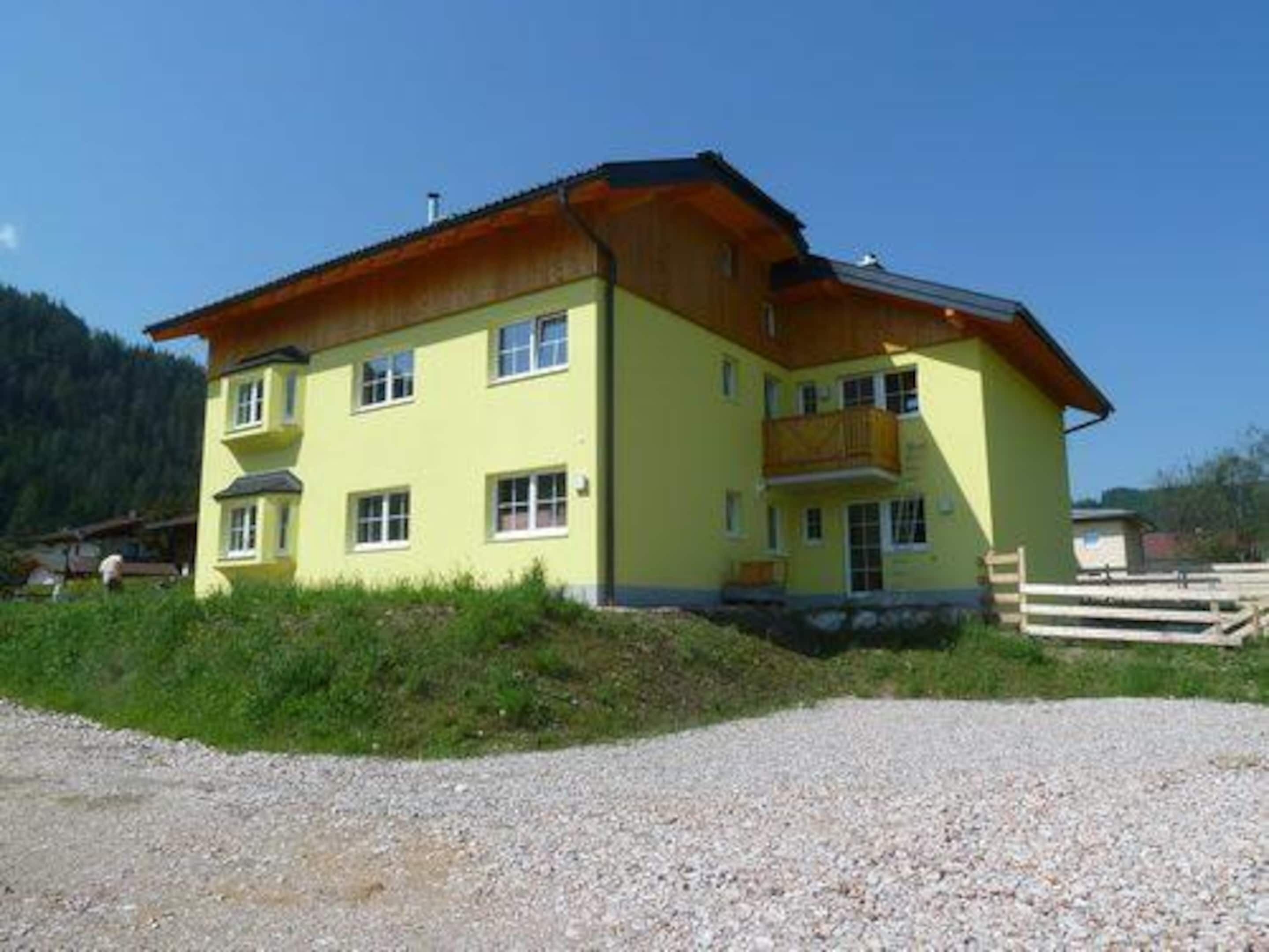 A two-story yellow building with a wooden balcony on the upper floor, surrounded by a gravel driveway and greenery.