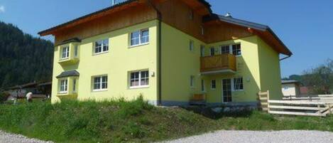 A two-story yellow building with a wooden balcony on the upper floor, surrounded by a gravel driveway and greenery.