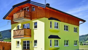 A multi-story yellow building with a wooden balcony, large windows, and a view of a green landscape. - Villa in Altenmarkt With a Balcony (Altenmarkt im Pongau)
