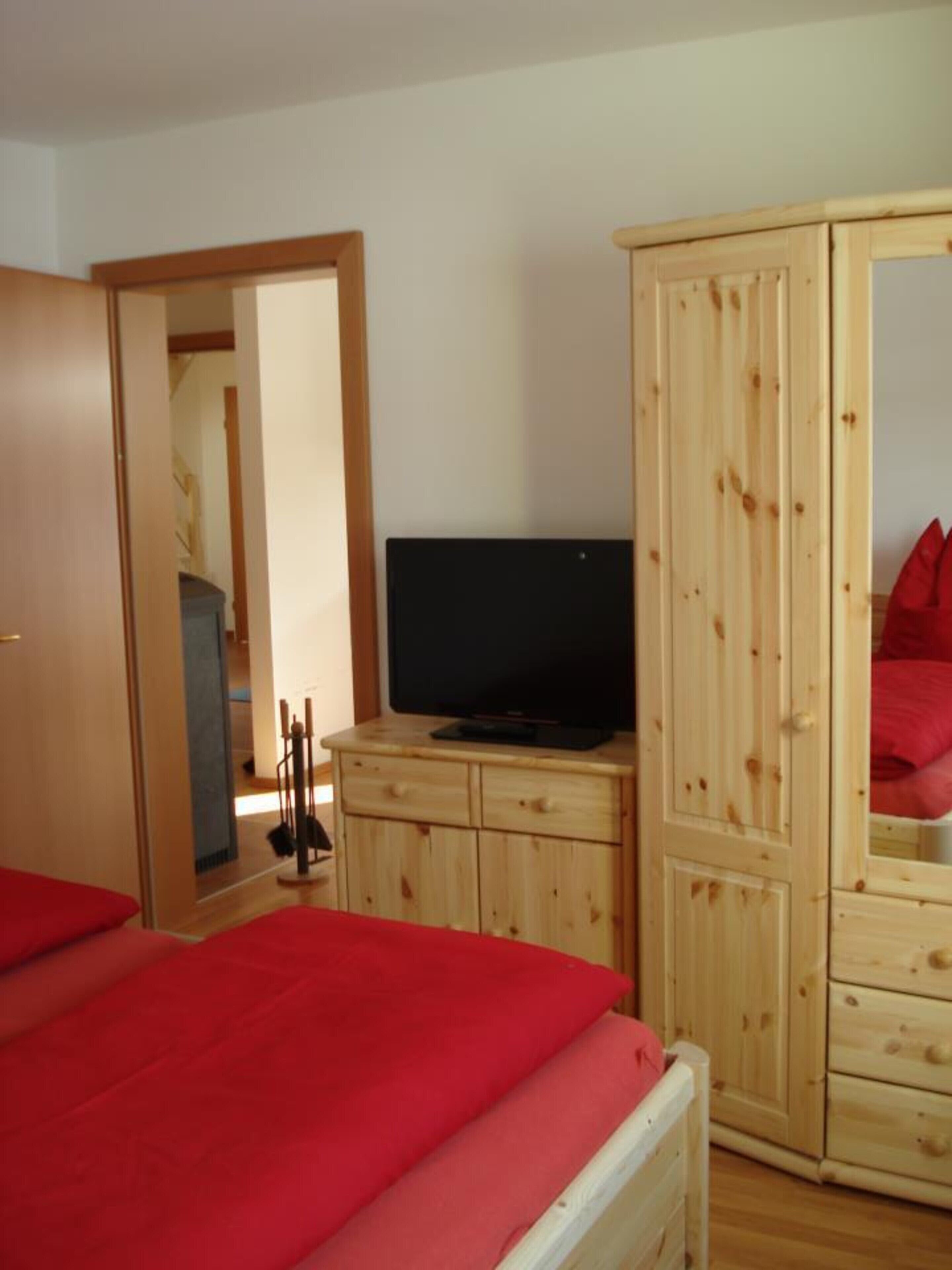 A bedroom with a wooden wardrobe, a television on top, and a red bedspread.