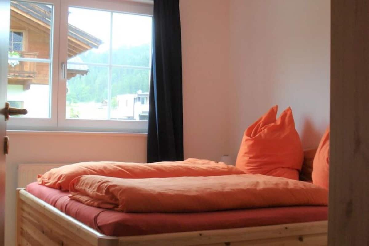 Un letto con biancheria arancione, una finestra vista su un edificio e montagne, e una testiera in legno.