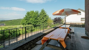 An outdoor patio with a table and benches, a stone wall, and a view of a forested area. - Spacious Holiday Home in Chodes With Sauna (Malmedy)