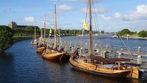 En række traditionelle træbåde ligger fortøjet ved en bro, med en klar himmel og en bygning i det fjerne.