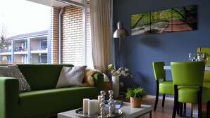 A modern living room with a green sofa, a coffee table with candles, a dining area with green chairs, and a large window with a view of a building. - Modern Apartment in Noordwijk Near the Sea (Noordwijk)
