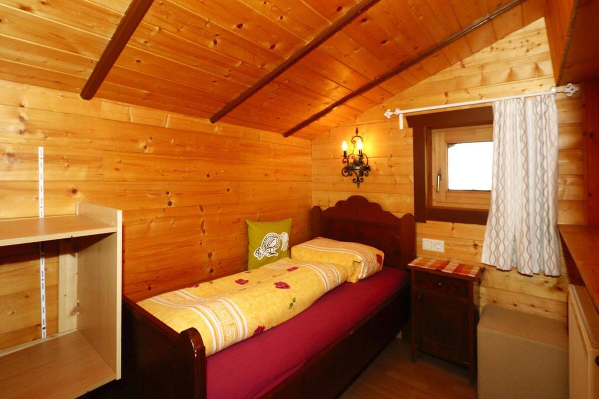A wooden cabin bedroom with a single bed, a nightstand, a wall-mounted lamp, and a window with curtains.