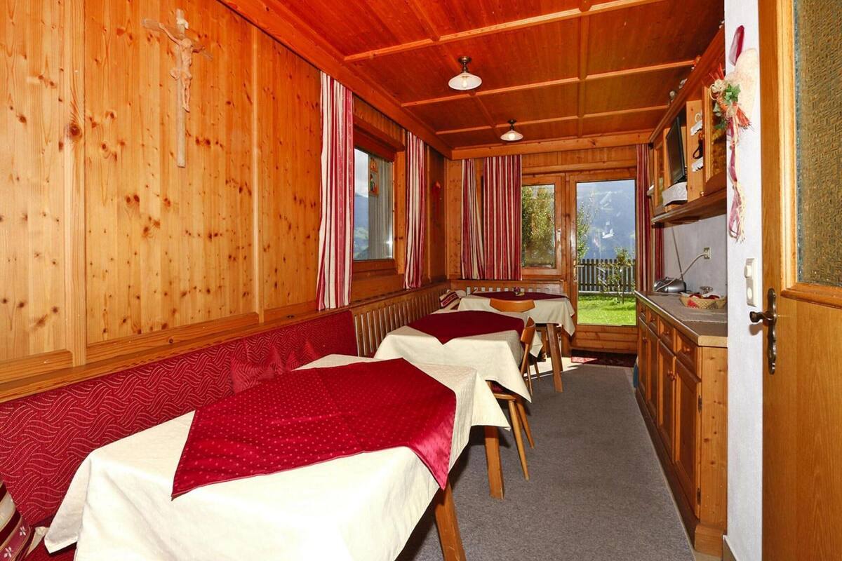 A cozy room with wooden walls and ceiling, featuring a long bench with beds, a small table, and a window with a view of the outdoors.
