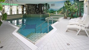 Una piscina interior con vistas a un mural de paisaje montañoso, rodeada de sillas reclinables blancas y un suelo de baldosas.