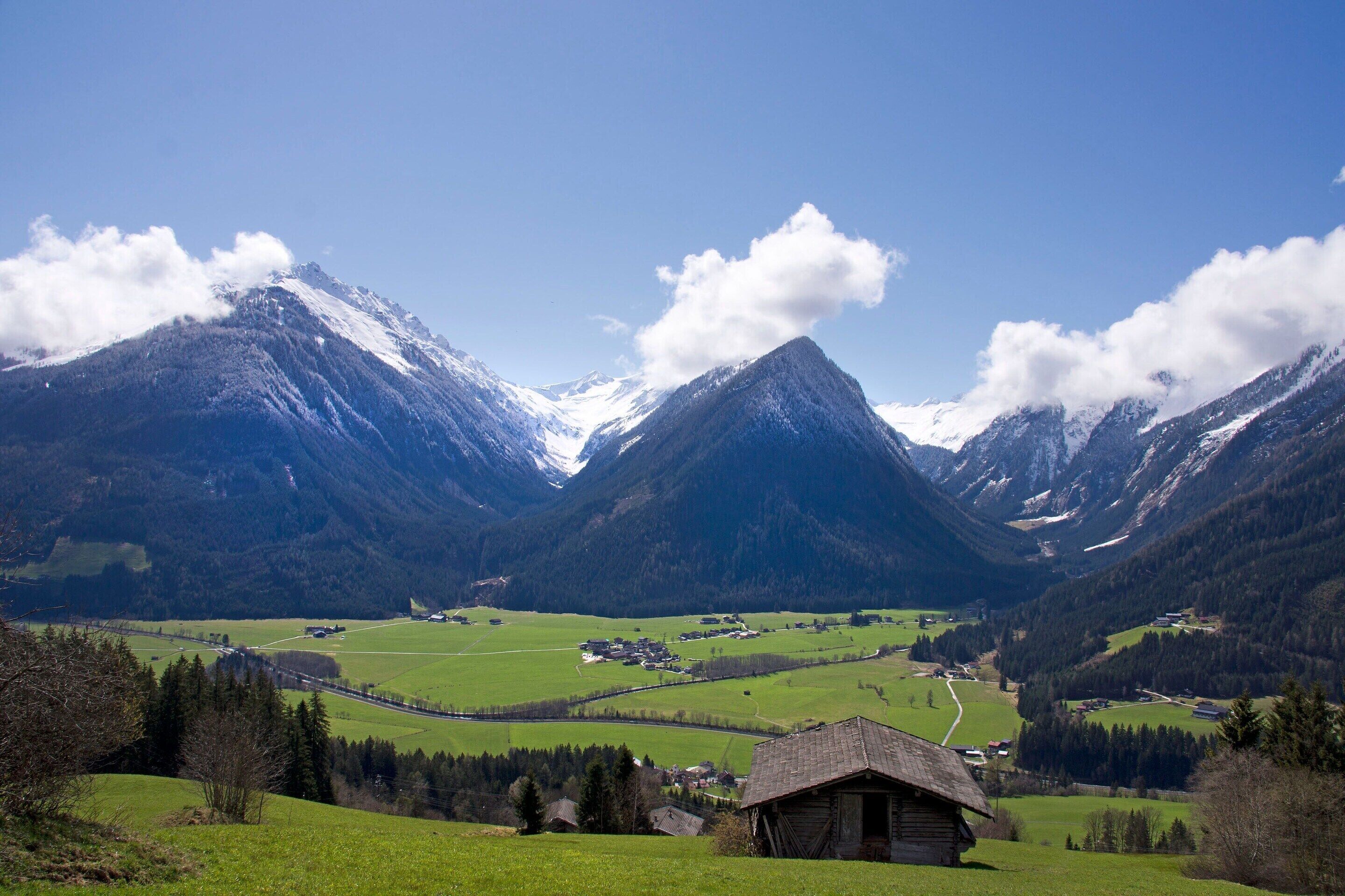 A mountain landscape with a small wooden cabin, a winding road, and snow-capped peaks.