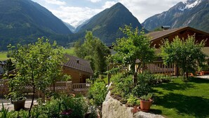 En bergsstuga med en stenmur och ett trästaket, omgiven av frodig grönska och en dal med berg i bakgrunden.