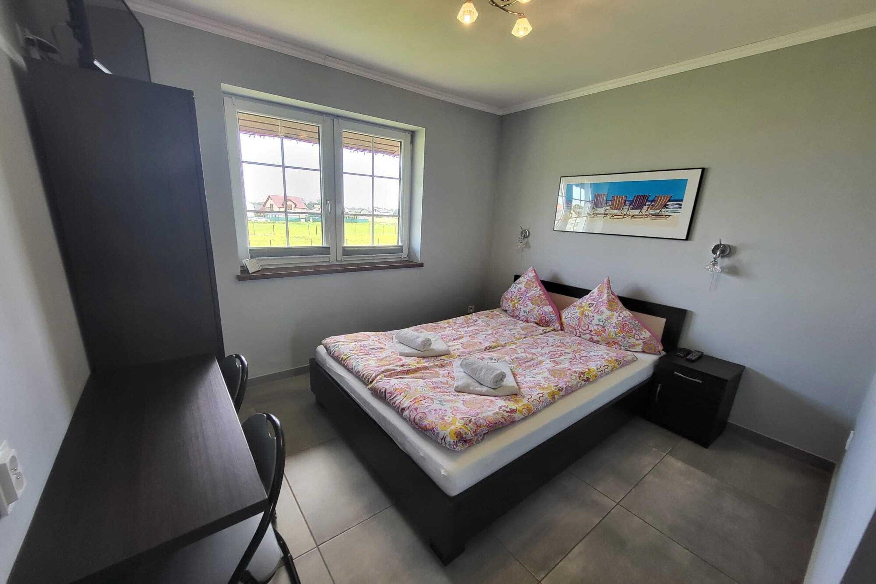 Een slaapkamer met een bed, een bureau, een stoel, een raam en een schilderij aan de muur.