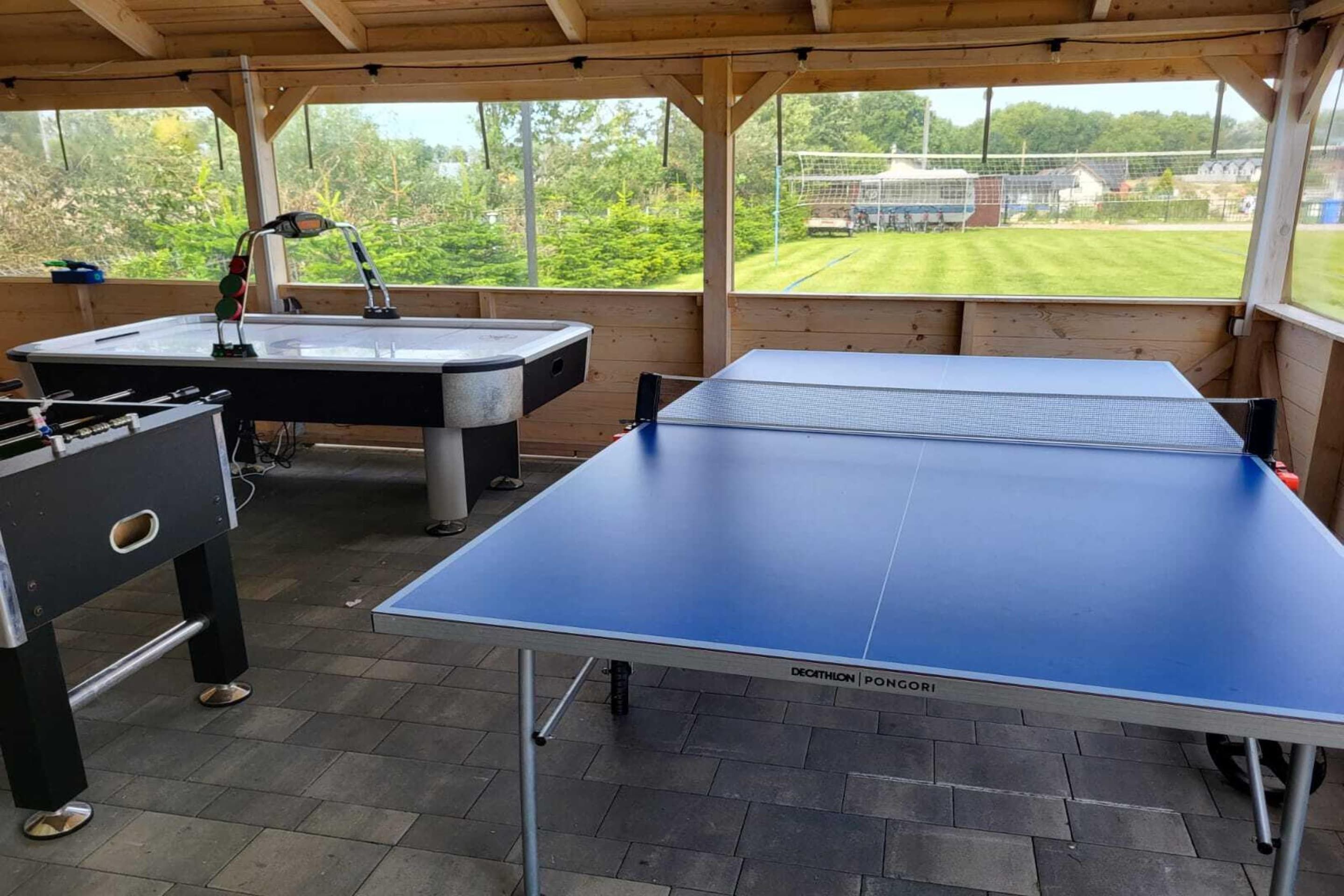 Et udendørs bordtennisbord med net, et fodboldbord og et ping pong bord under et træbeklædt pavillon med udsigt over en græsplæne.