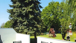 Een terras met een tafel en stoelen, een houten speeltoestel en een grote boom.