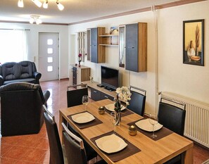 Uma área de refeições com mesa de madeira posta para quatro pessoas, um sofá e uma TV fixada na parede.