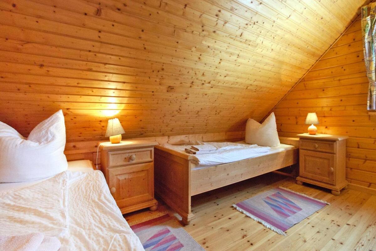 A cozy attic bedroom with wooden walls, a single bed, two bedside tables with lamps, and a colorful rug.