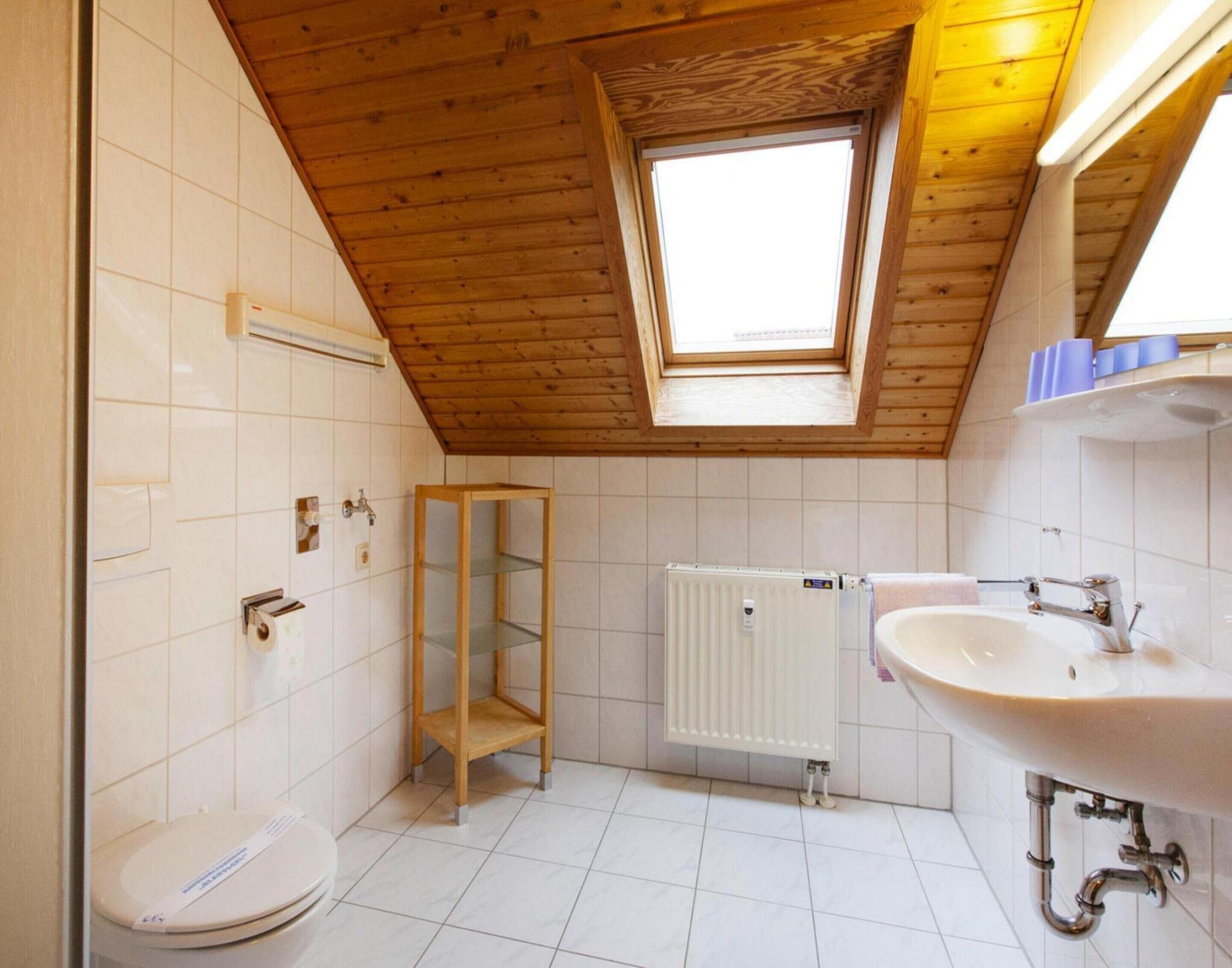 A small, bright bathroom with a skylight, white tiles, a toilet, a wooden shelf, and a radiator.
