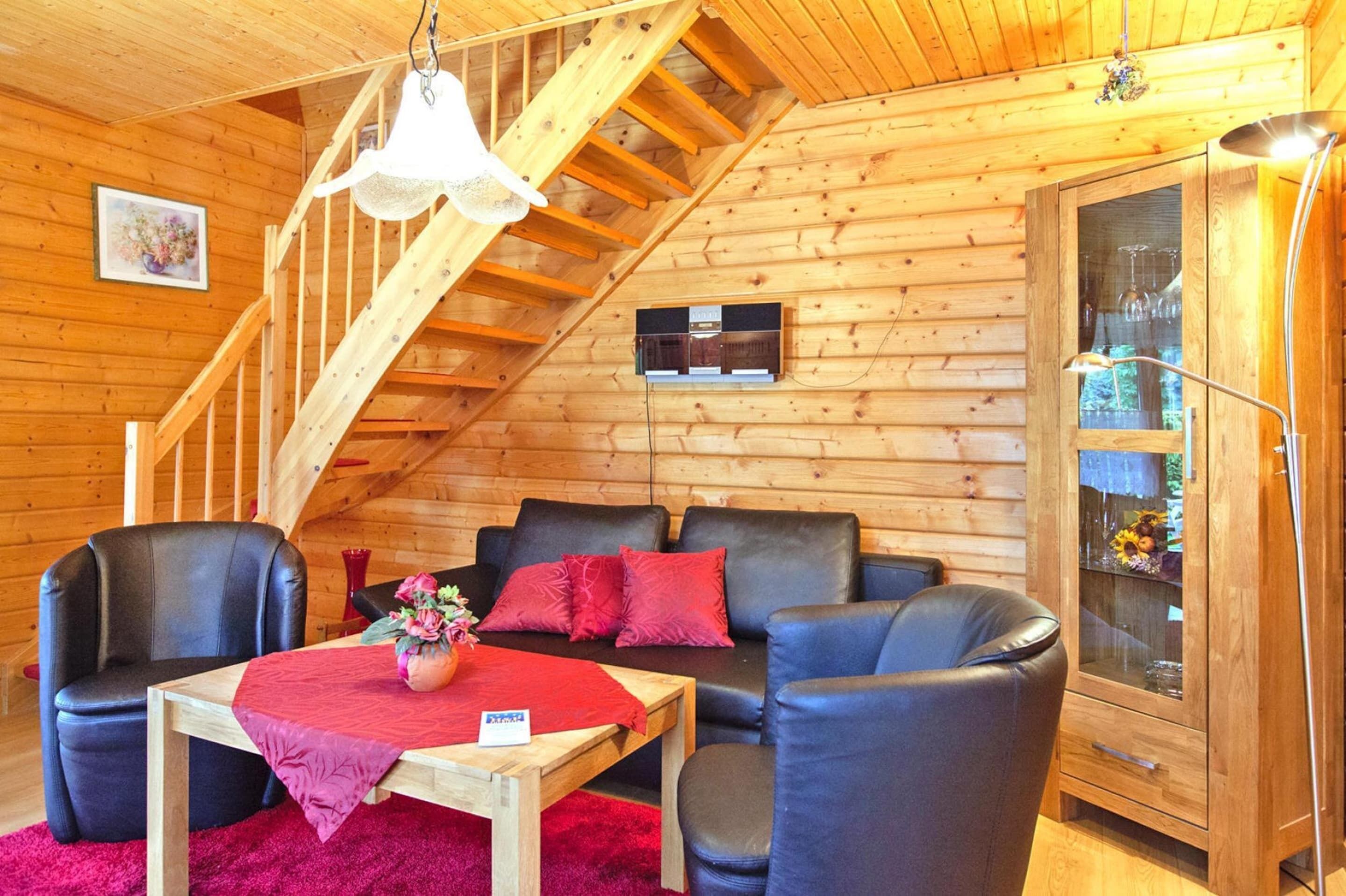 A cozy log cabin interior with a wooden staircase, a sofa set, a table with a red tablecloth, and a cabinet with glass doors.