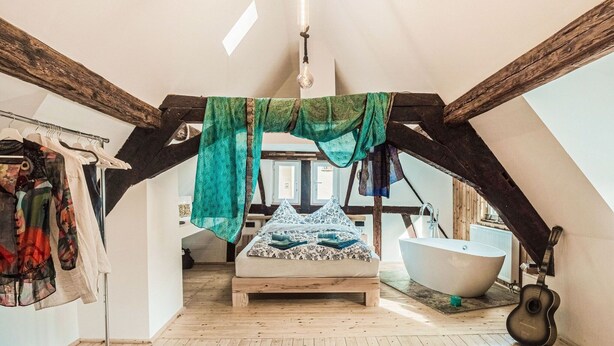 A loft bedroom with a sloped ceiling, a wooden bed, a large window, a bathtub, and a guitar. - Cosy Holiday Home in Sankt Goarshausen With Barbecue (Sankt Goarshausen)