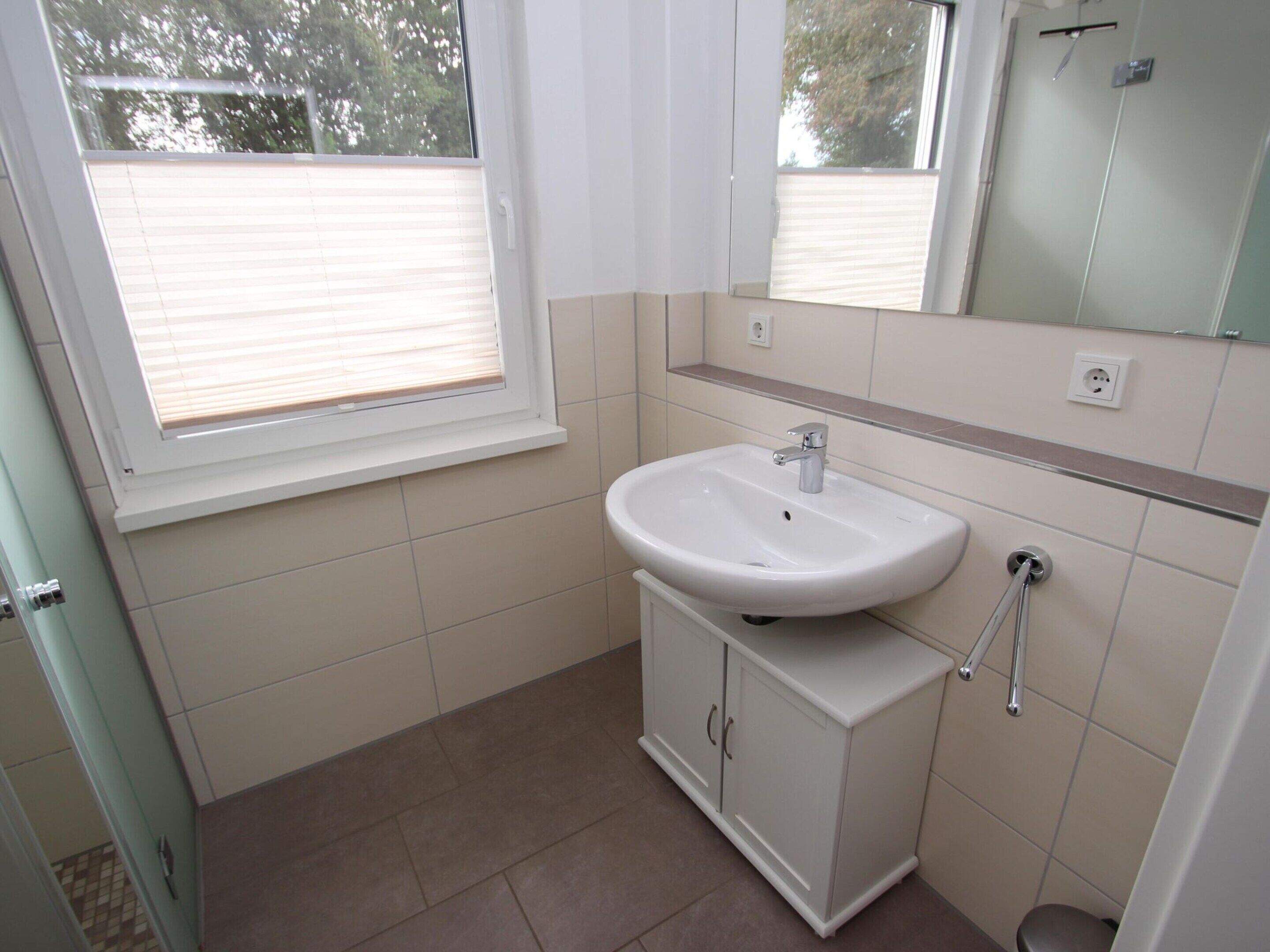 A bathroom with a white sink, cabinet, and mirror. There is a window with blinds, and the floor is tiled.