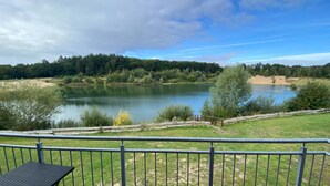 A lakeside view from a deck with a metal railing, a small table, and a grassy area in the foreground. - Ferienhaeuser im Ferienpark, Hechthausen (Hechthausen)