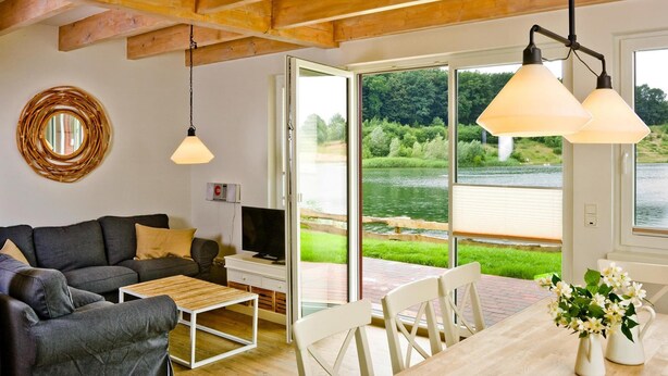 A modern living room with a sofa, coffee table, and dining table. There is a TV on a stand, a wicker wall decoration, and a view of a lake through large glass doors. - Ferienhaeuser im Ferienpark, Hechthausen (Hechthausen)