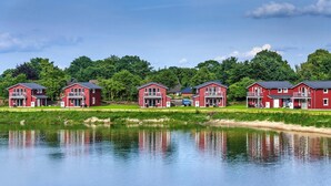 A row of red buildings with balconies, a pond in the foreground, and trees in the background. - Ferienhaeuser im Ferienpark, Hechthausen (Hechthausen)