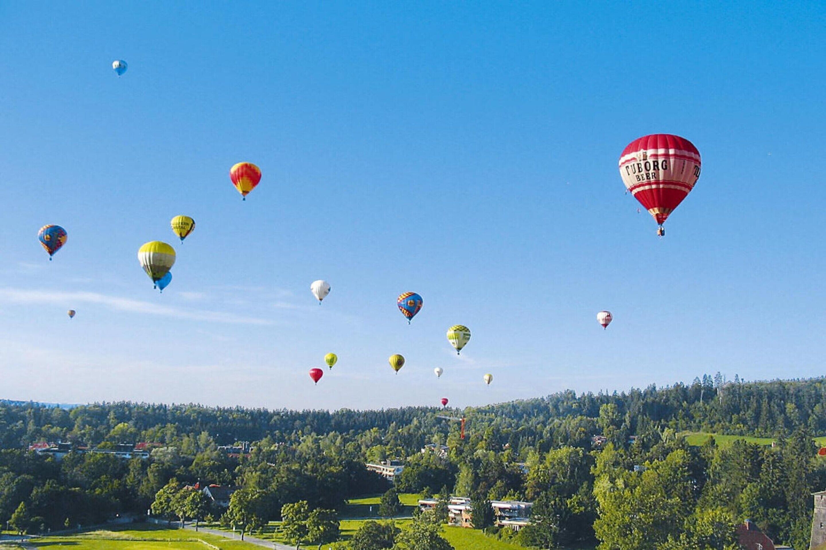 A clear sky with multiple hot air balloons of various colors floating above a landscape with houses and trees.