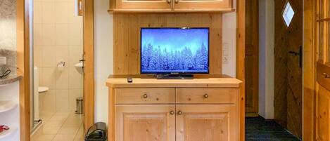 Ein Holzkabinet mit einem Fernseher, der eine verschneite Waldszene zeigt, ein Mülleimer sowie ein Badezimmer mit Toilette und Waschbecken.