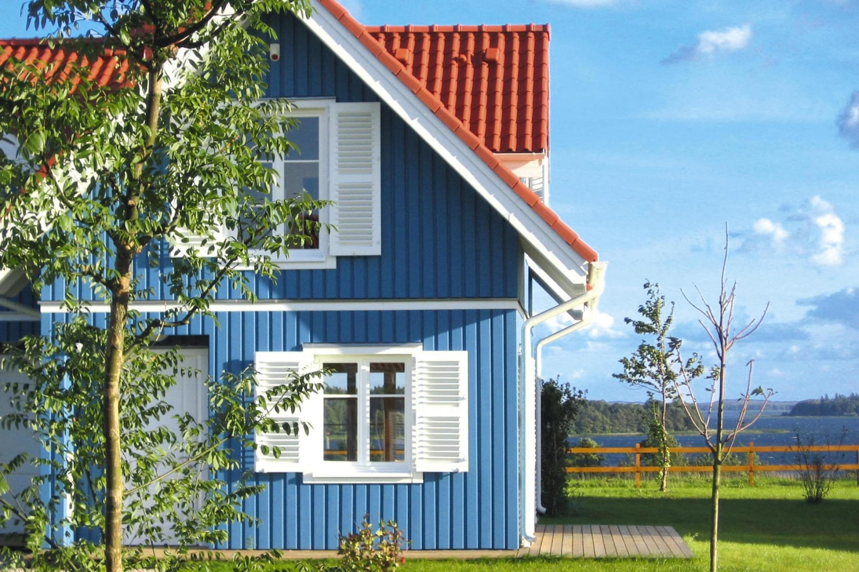 A blue house with white shutters, a red roof, and a view of a lake.