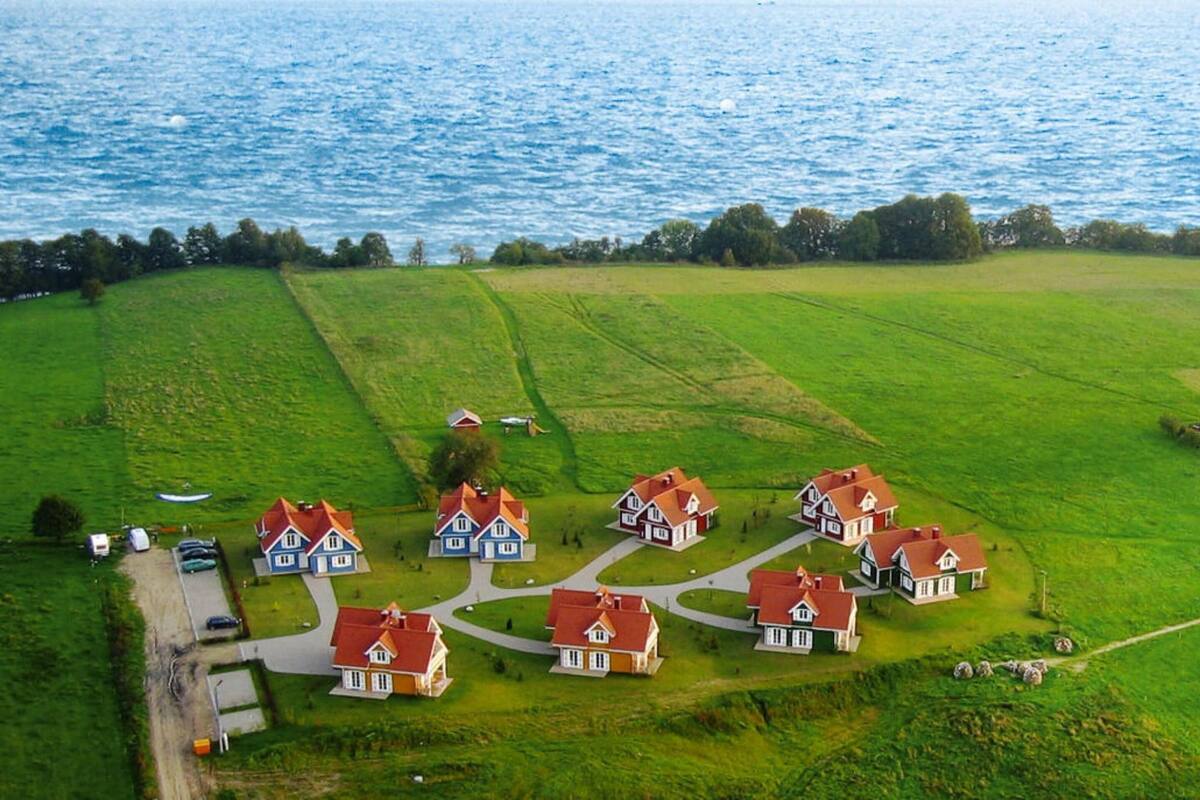 An aerial view of a coastal village with houses arranged in a circular pattern around a central green space, with a road leading to the sea.