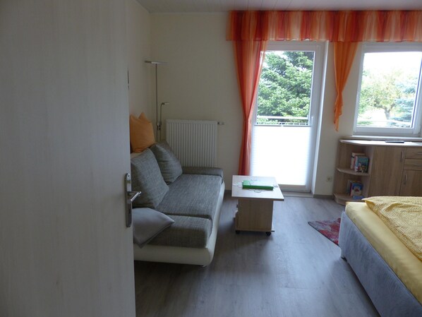 A compact room with a sofa, a small table, a bed, and a window with curtains.