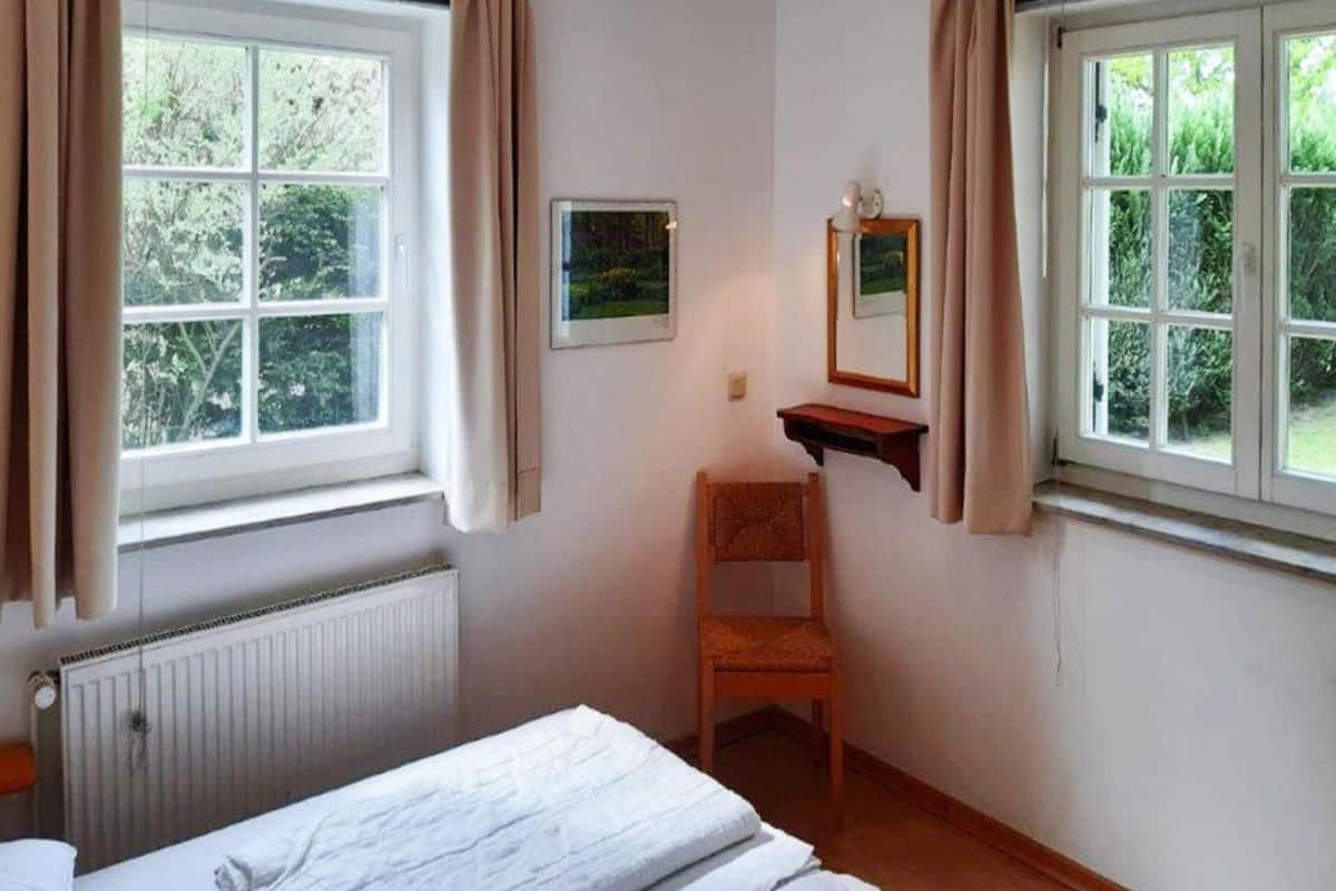 A small, tidy room with a single bed, a chair, and a small table. There are two windows with curtains, and a radiator is visible on the wall.