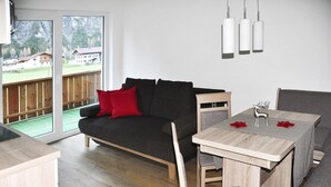 A modern living room with a dark grey sofa, red cushions, a wooden dining table with grey chairs, and a view of a wooden deck and mountains through large windows. - Apartment in Steeg in a Beautiful Setting (Steeg)