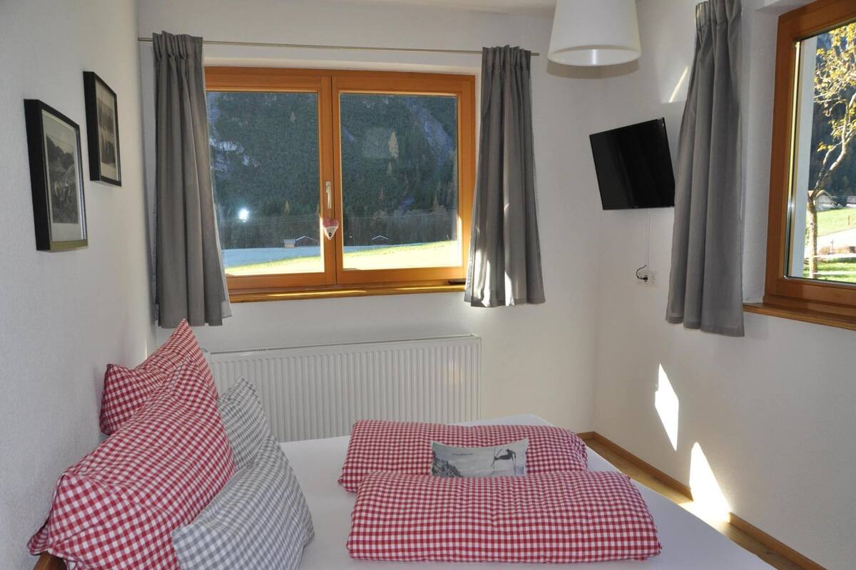 A bedroom with a bed, checkered bedding, a window with curtains, a mounted TV, and framed pictures on the wall.