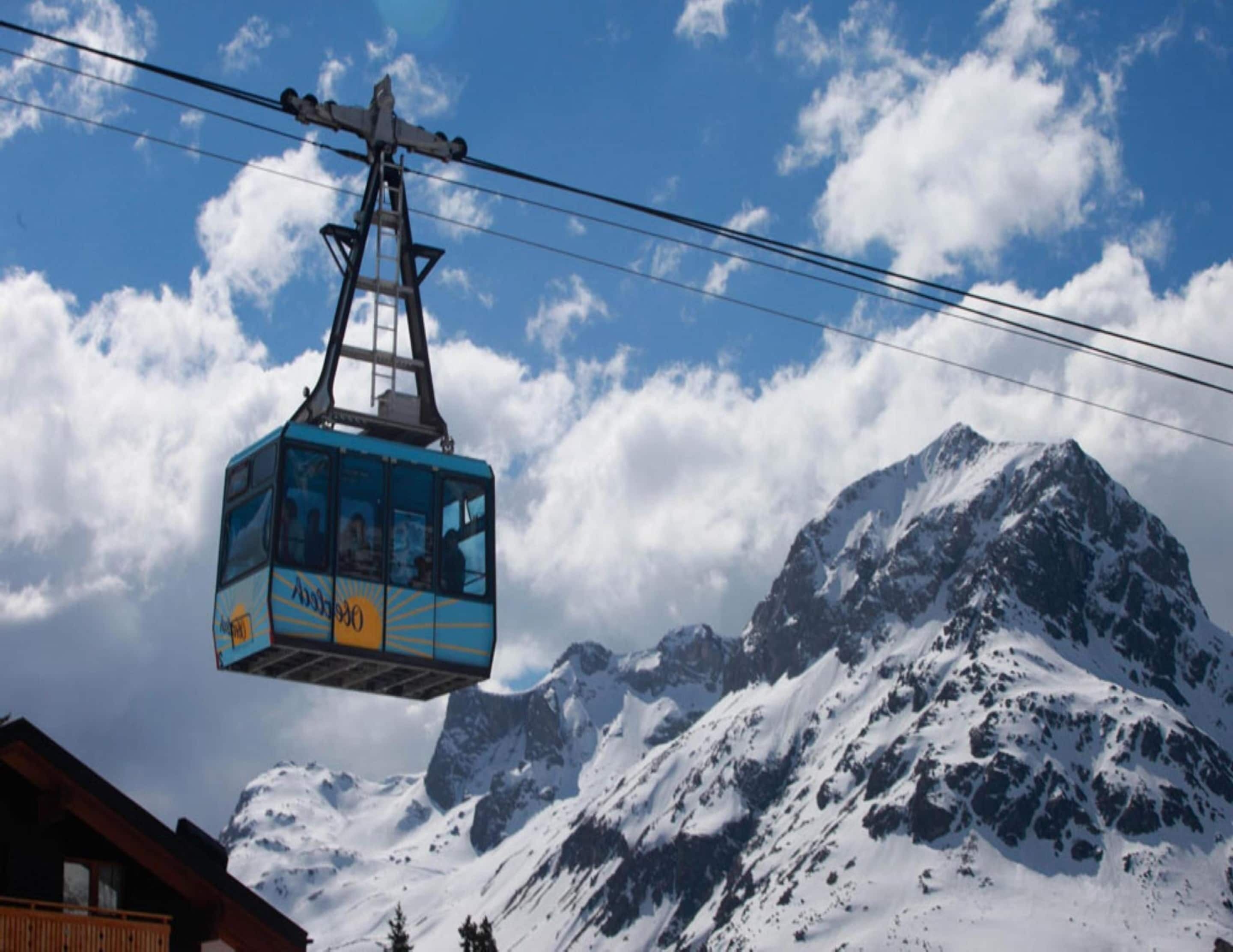 A cable car with passengers, suspended by cables against a backdrop of snow-covered mountains and a partly cloudy sky.