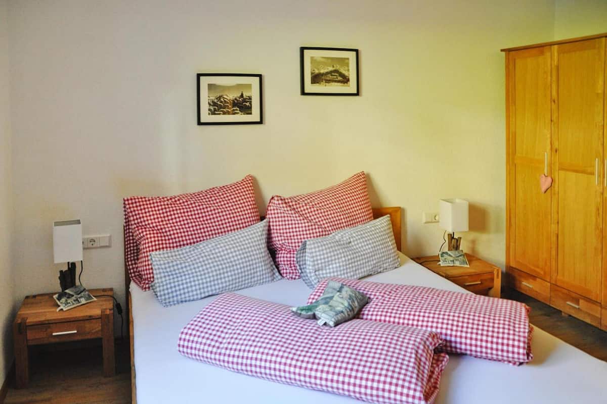 A neatly made bed with checkered bedding, a bedside table with a lamp, a wooden wardrobe, and two framed pictures on the wall.