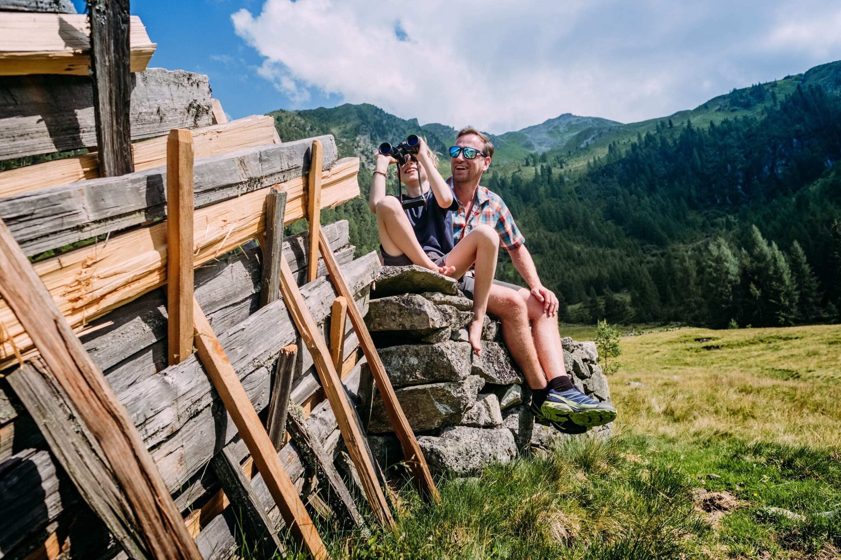 Two people sitting on a wooden bench with a mountainous landscape in the background.