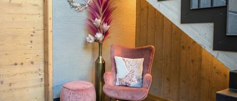 Un interior moderno con una pared de madera, un sillón tapizado rosa con un cojín decorativo, un banquillo redondo pequeño y un jarrón con flores secas.