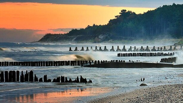A coastal scene with a rocky shoreline, waves crashing against a breakwater, and a forested hill in the background. - Holiday Home in Jaroslawiec With a Terrace (Postomino)