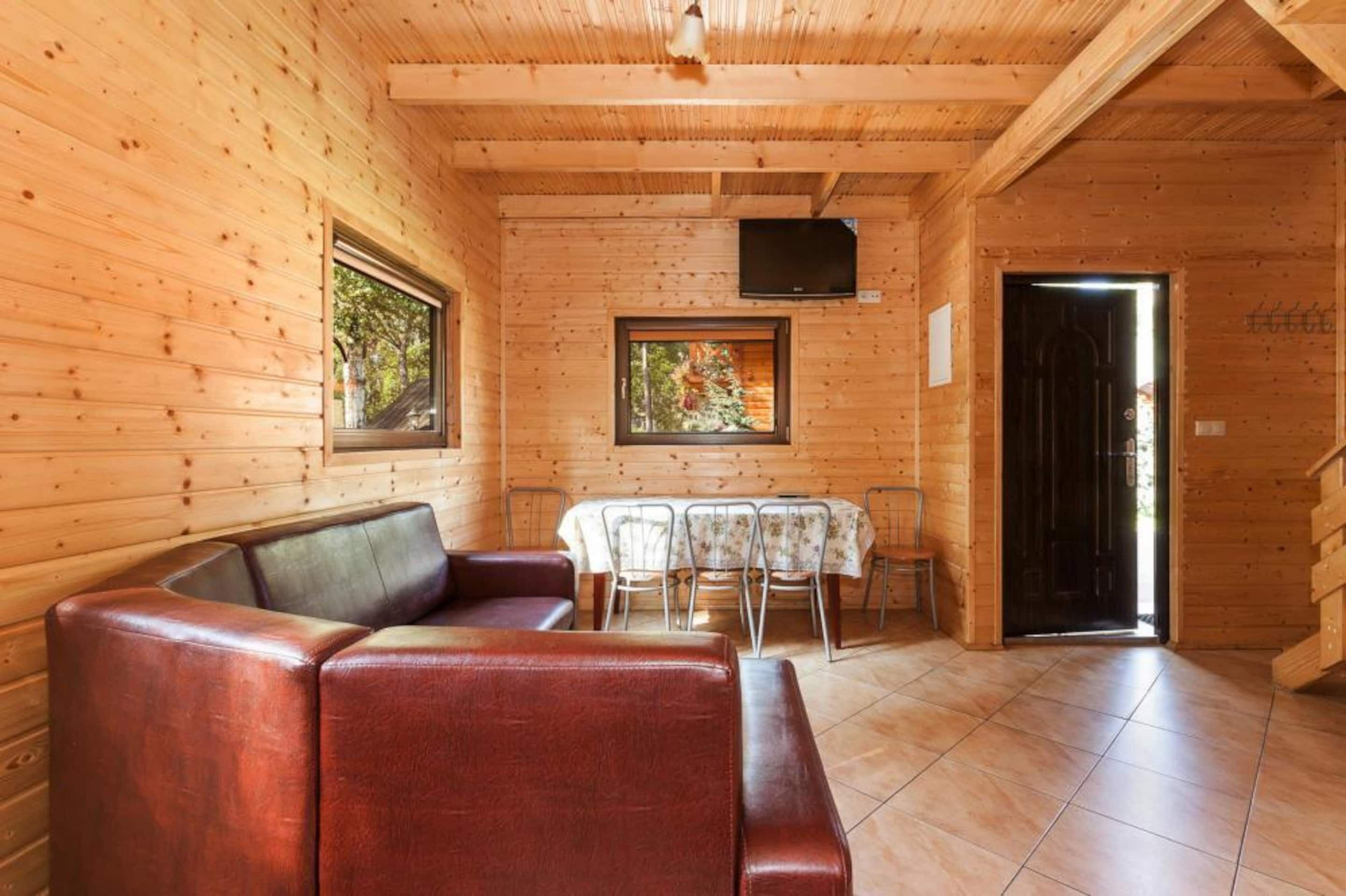 A wooden cabin interior with a leather sofa, dining table, chairs, and a TV mounted on the wall.