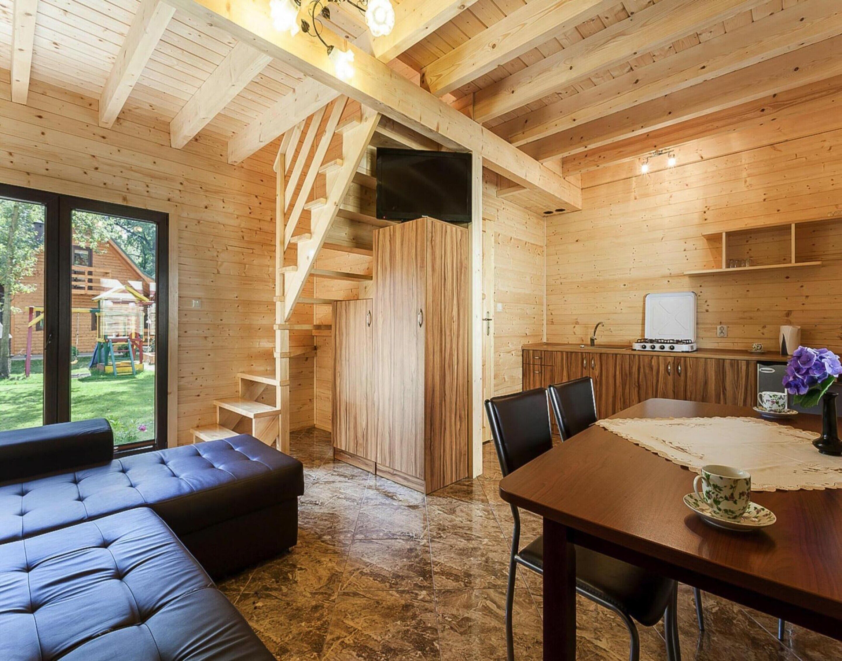 A wooden cabin interior with a sofa, dining table, chairs, a kitchen area, and a staircase leading to a loft bedroom.