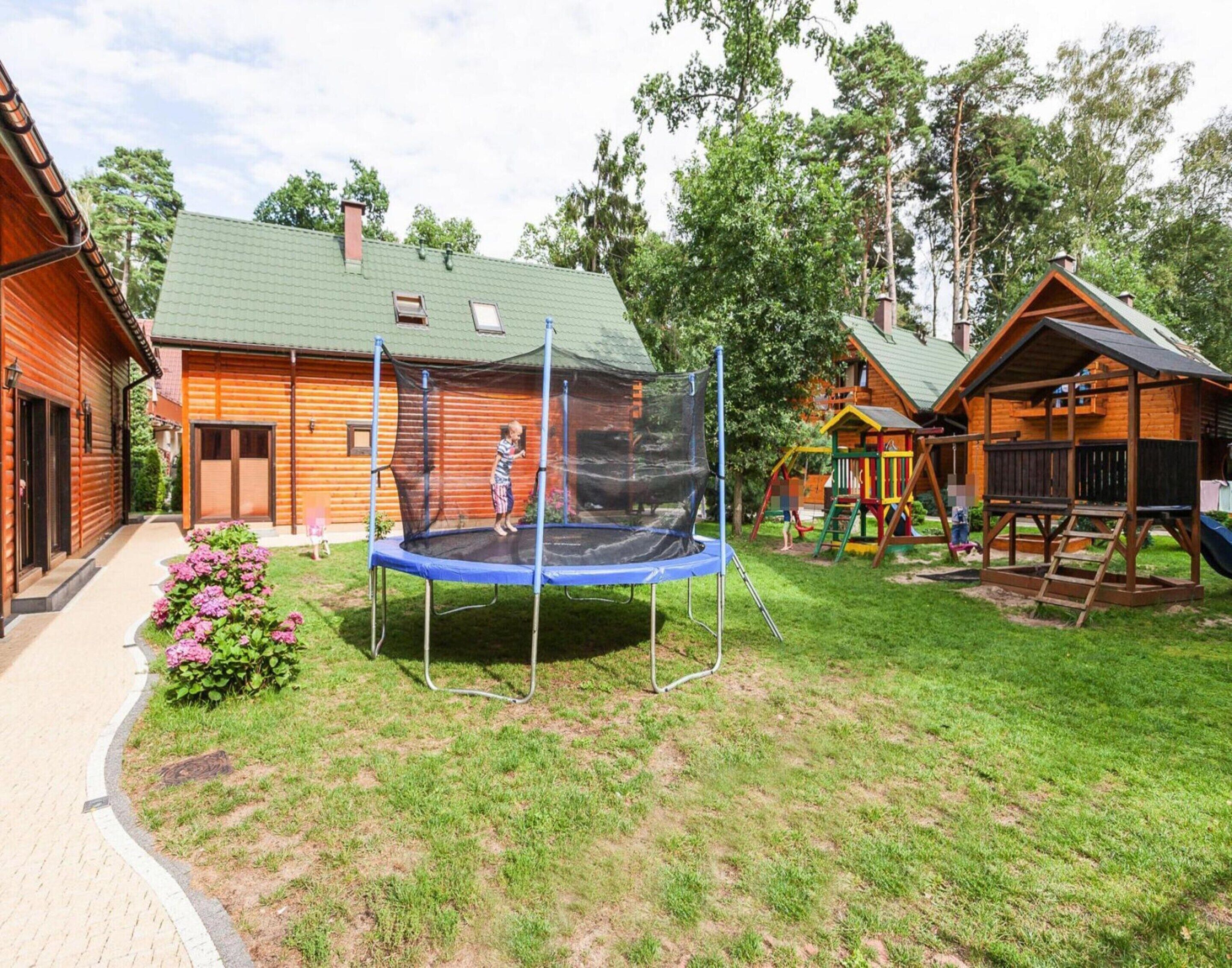 A wooden cabin with a trampoline in the yard, surrounded by trees and a small playground.