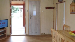 Una habitación con piso de madera, un televisor sobre un mueble bajo, una mesa de comedor con sillas y una puerta que da a un espacio exterior.
