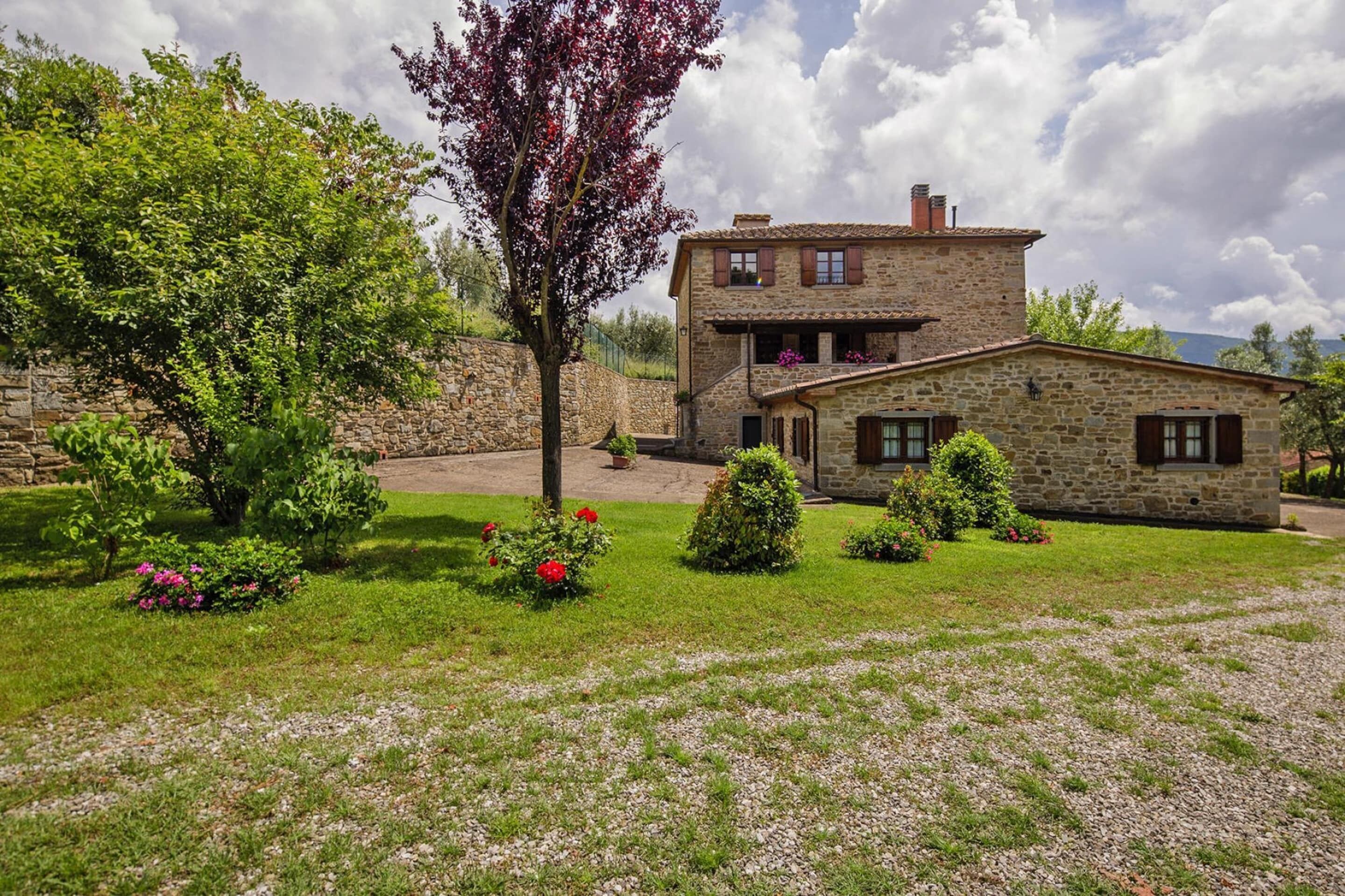 A stone house with a balcony, surrounded by a well-maintained garden with various shrubs and flowers.