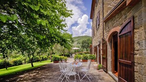 A stone building with a paved courtyard, a table with chairs, and a large tree. - Farmhouse Le Capanne, Castiglion Fiorentino (Castiglion Fiorentino)