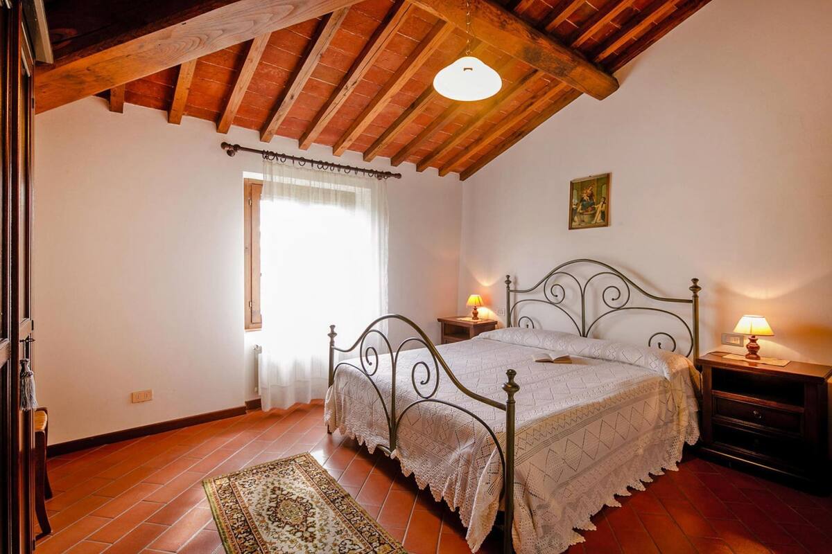 A bedroom with a wooden ceiling, a bed with a metal frame, a nightstand with a lamp, and a window with a curtain.
