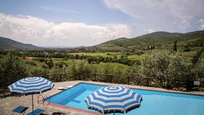 A pool with two striped umbrellas, surrounded by a stone deck and a wooden fence, overlooking a lush valley with hills and trees. - Farmhouse Le Capanne, Castiglion Fiorentino (Castiglion Fiorentino)