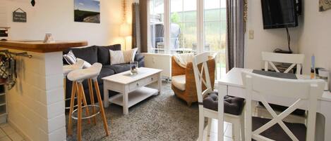 A compact living space with a sofa, coffee table, and dining area. A TV is mounted on the wall, and there is a window with curtains.