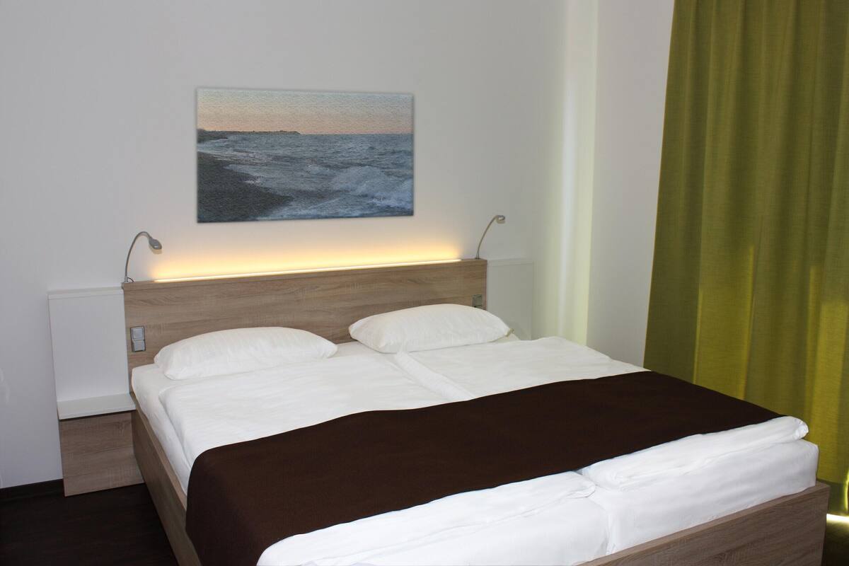 A neatly made bed with white linens and a brown blanket, a headboard with a picture of the sea, and a green curtain to the right.